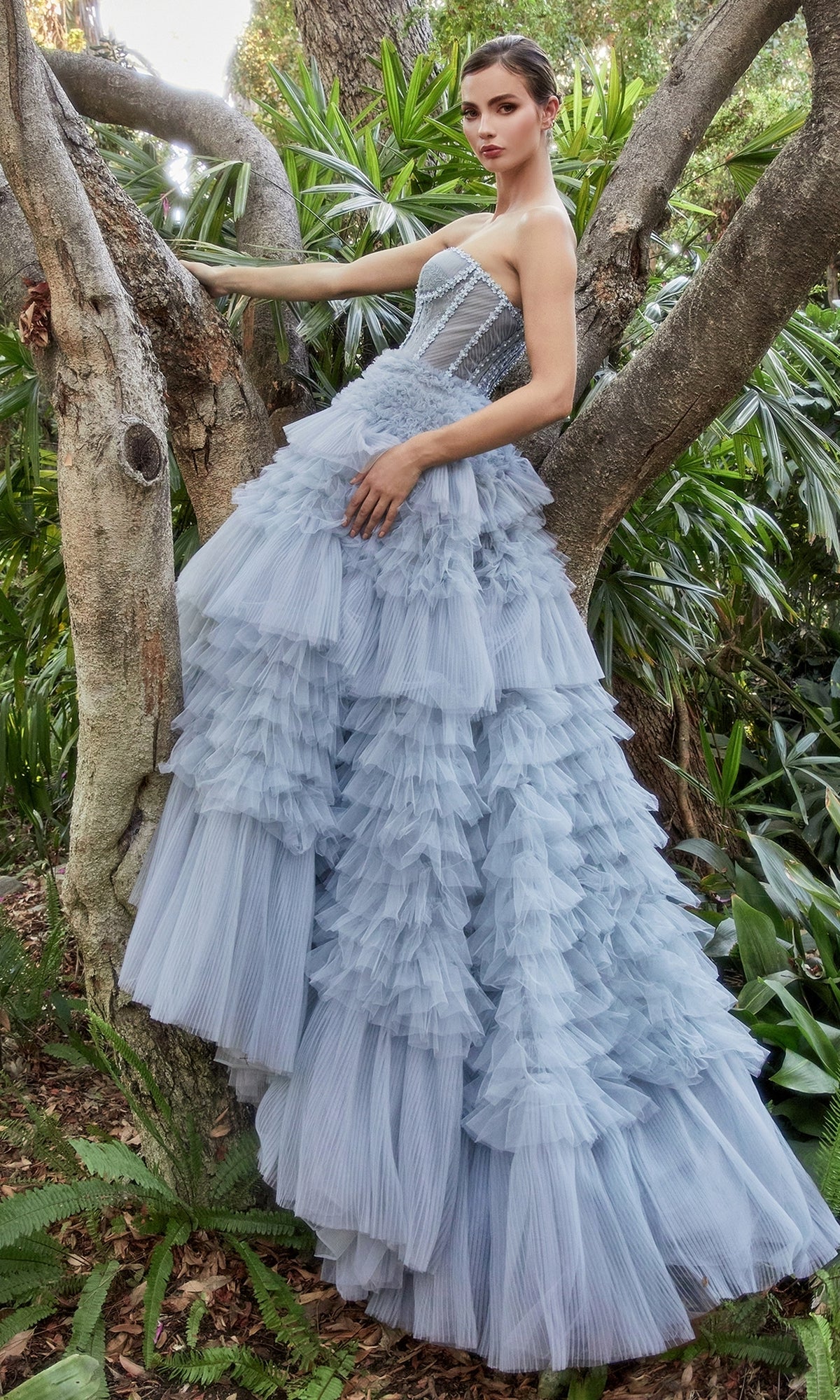 A woman in the Strapless Tiered Long Prom Ball Gown A1017 with ruffled tulle stands by a tree in a lush, green outdoor setting.